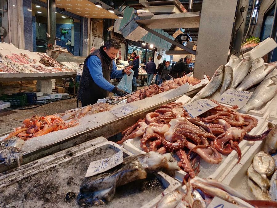 A man stands in a bustling fish market, wearing gloves, as he carefully picks through a pile of freshly caught fish, selecting the best ones to take home. The bright colors and variety of fish create a vivid and lively scene.
